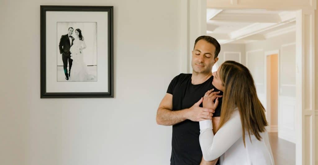 A woman embracing a man in a black shirt, with a framed picture of a couple on the wall behind them.
