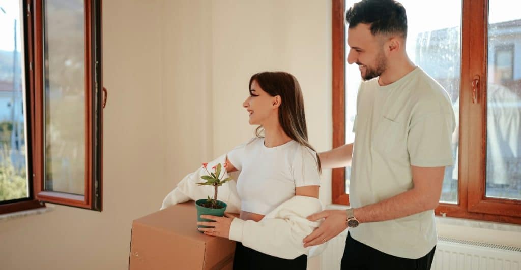 A man and woman walking in a room with light-colored walls, the woman carrying a small plant and the man carrying a box, both smiling.