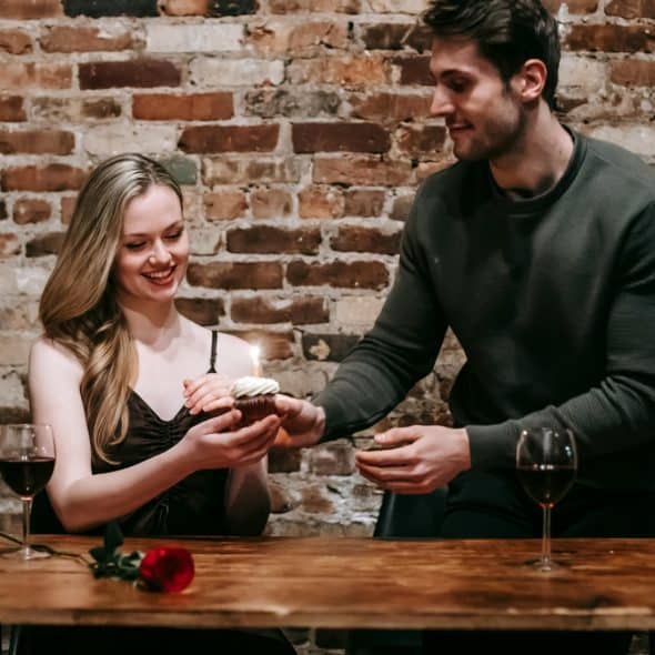 A man giving a woman a cupcake with a lit candle at a table with wine glasses and a rose, in front of a brick wall.