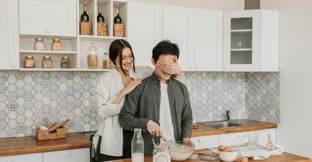 A woman covering a man's eyes from behind while he is mixing something in a bowl in a kitchen.