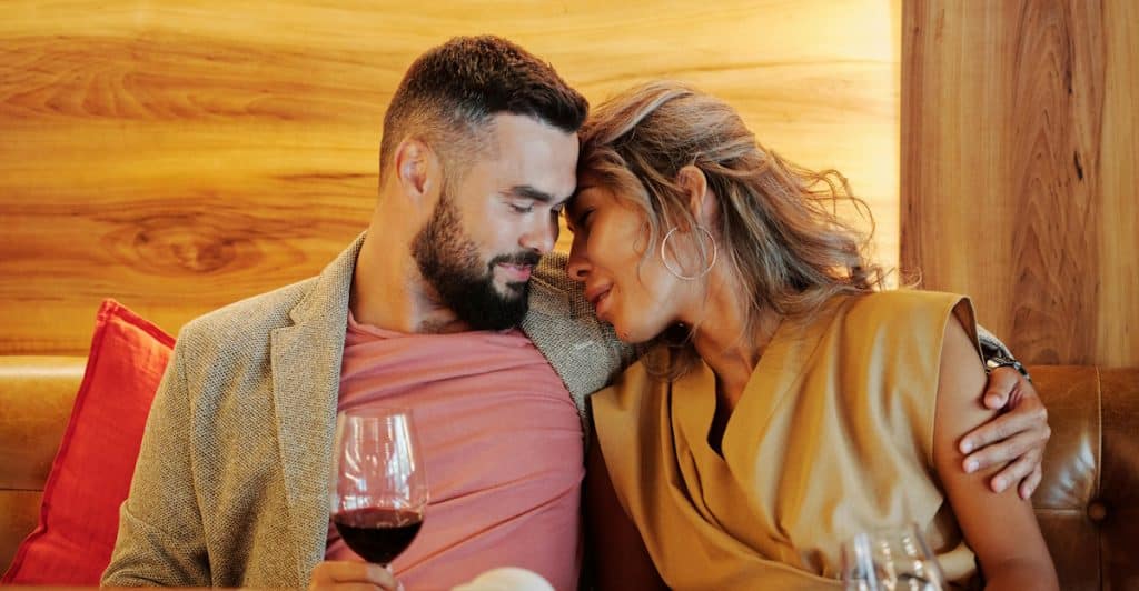 A man and a woman embracing at a table with drinks and phones, with the woman resting her head on the man's shoulder.