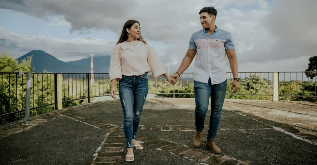 A man and a woman holding hands and walking on a paved area with mountains in the background.
