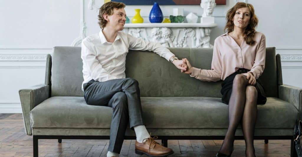 A man and a woman sitting on a grey couch, holding hands and looking in opposite directions.