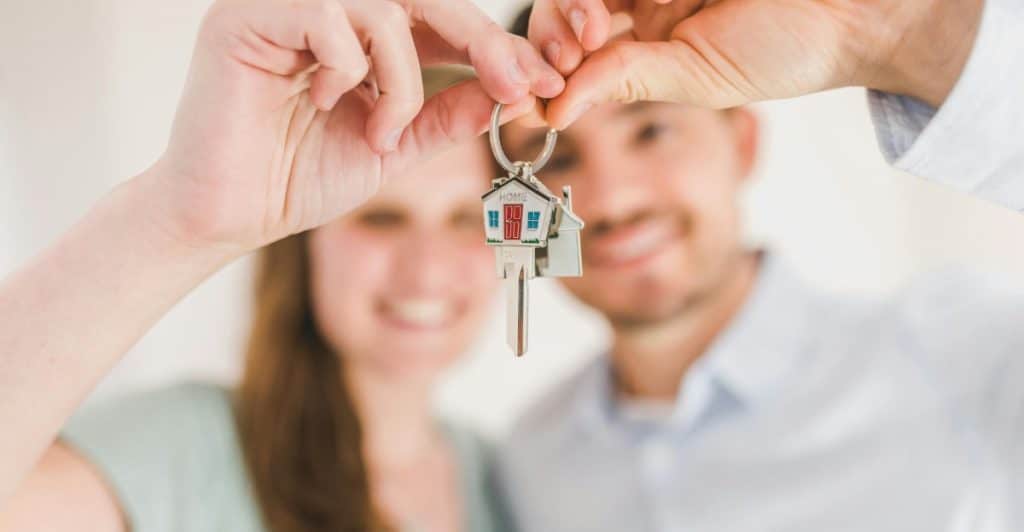 A blurry image of a smiling couple in the background, with their hands in the foreground holding up a set of house keys with a house-shaped keychain.
