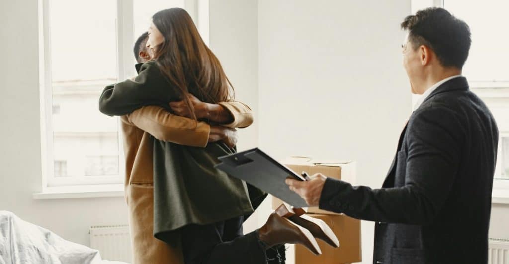 A man and a woman hugging in a room with cardboard boxes, while another person in a suit stands nearby holding a clipboard and looking at them.