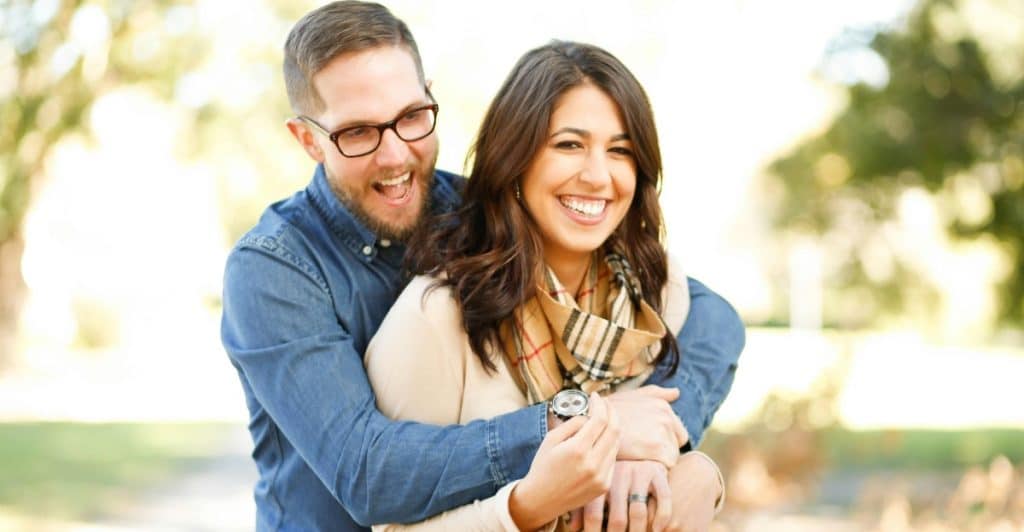  A man with glasses hugging a woman from behind in an outdoor setting, with both smiling.