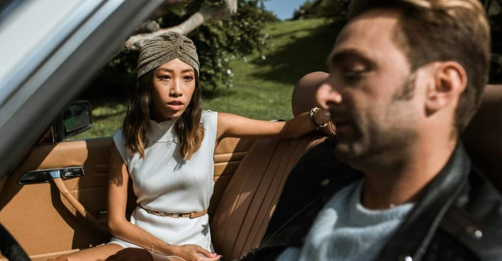 A woman in a head wrap looking at a man who is looking away while sitting in a car.