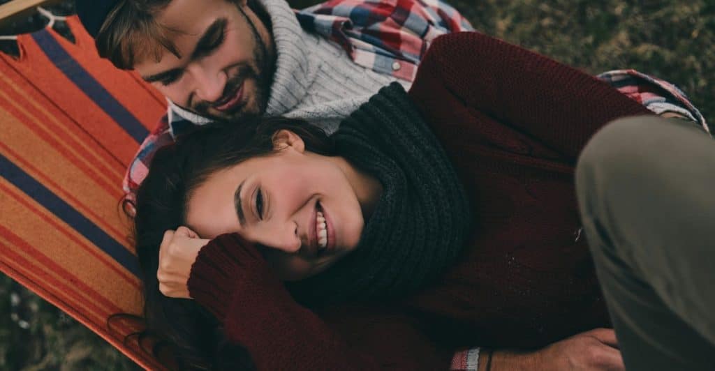 A man is lying in a hammock with his arm around a smiling woman who is resting her head on his chest.