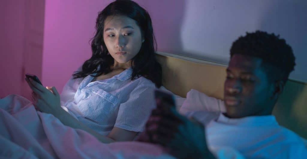 A person with light skin on the left and a person with dark skin on the right lying in bed, both looking at their phones with the room lit by purple and blue light