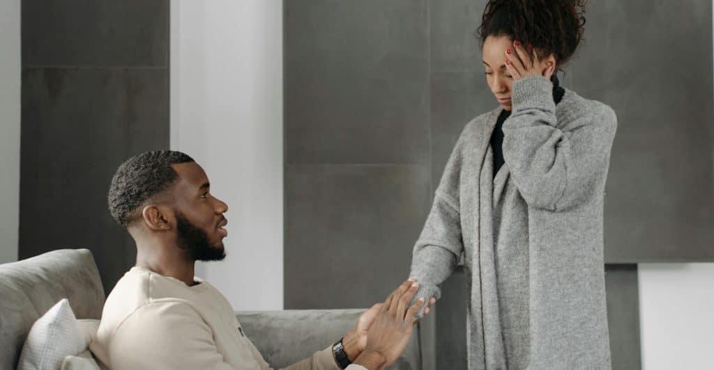 A man sitting on a couch holding the hand of a woman who is standing and touching her forehead, appearing distressed.