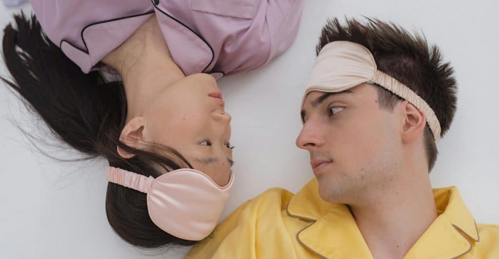 A man and a woman lying on a white surface, both wearing eye masks and looking at each other.