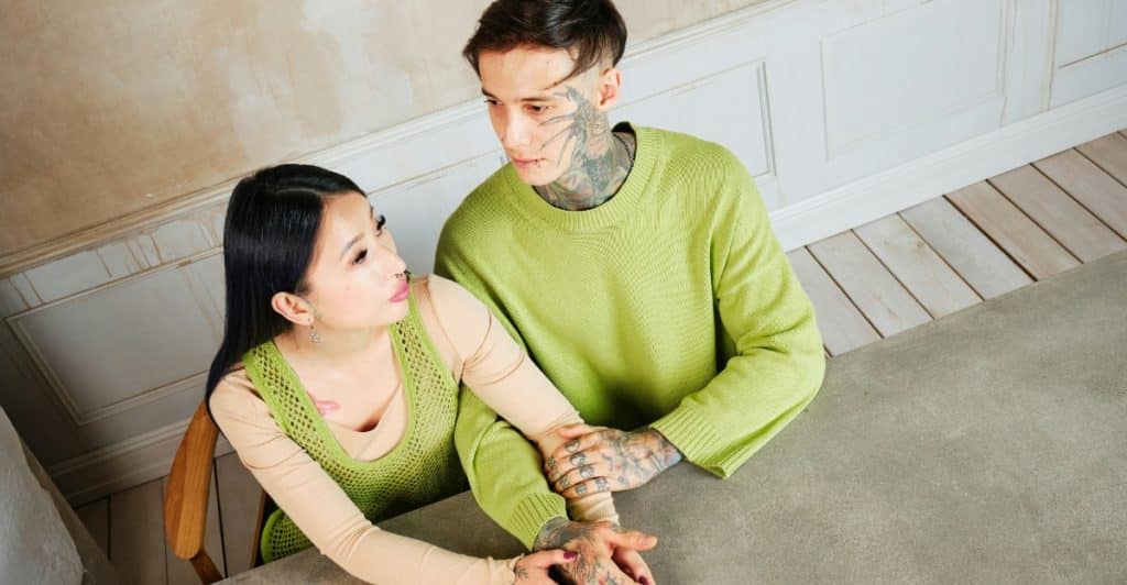 A man with tattoos on his neck and face is sitting at a table, holding the hands of a woman with long black hair who is looking at him.