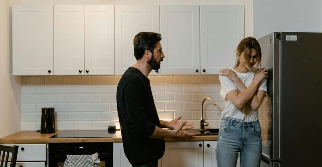A man and a woman standing in a kitchen, with the man talking to the woman who is leaning against a fridge.