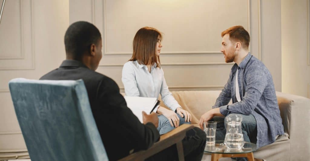 A couple sitting on a couch across from a therapist, who is seated in a chair and taking notes.