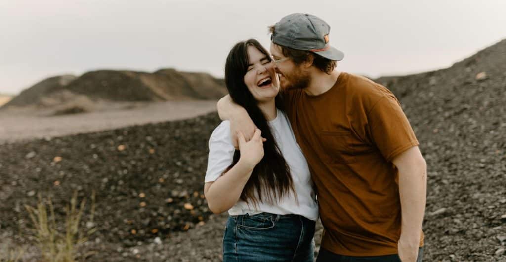 A man with a beard is embracing a woman from behind, and both are laughing with a pile of rocks in the background.