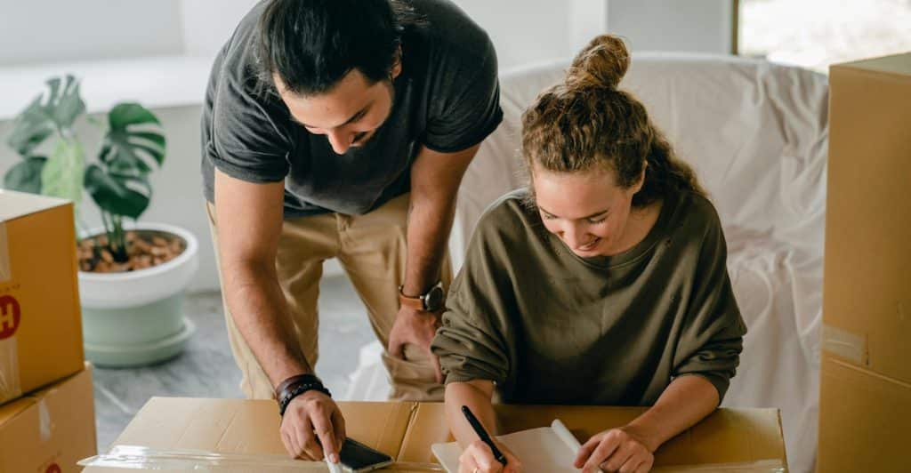 A man and a woman looking down at a large cardboard box, with the woman holding a pen and notebook.