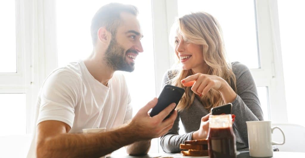 A man and a woman sitting at a table, smiling and looking at a smartphone together.