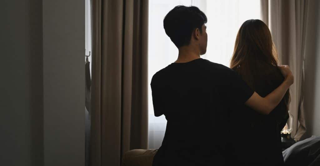 A couple sitting on a bed, looking out a window with curtains, their backs to the viewer