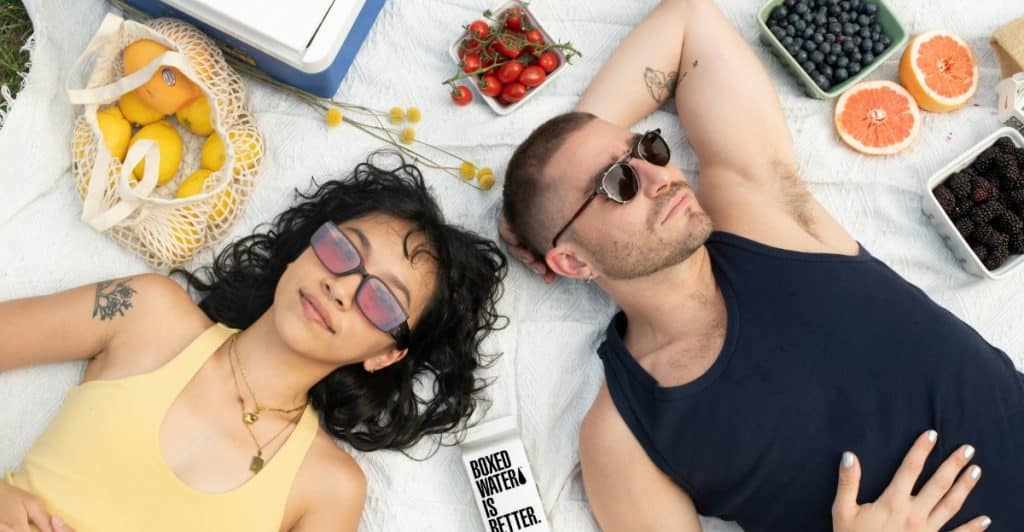 A man and a woman lying on a blanket outdoors, surrounded by picnic food like fruit, with sunglasses on.