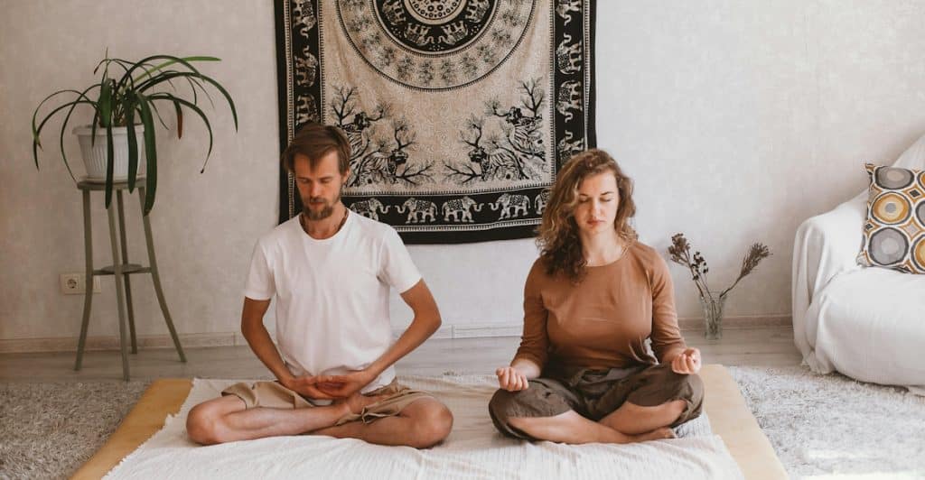 A man and a woman sitting cross-legged on mats in a room, with their eyes closed, in a meditative pose.