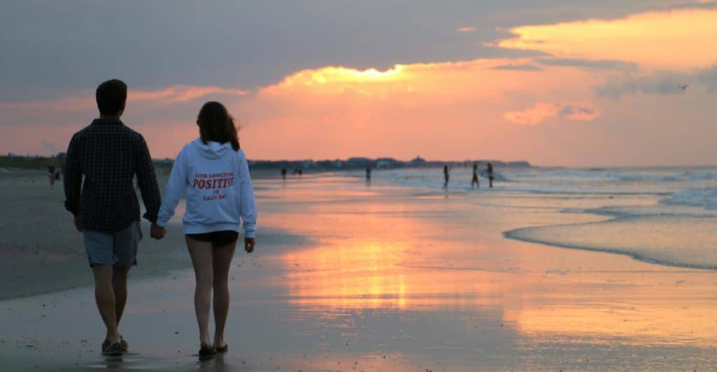 A man and a woman holding hands and walking away from the camera on a beach at sunset.