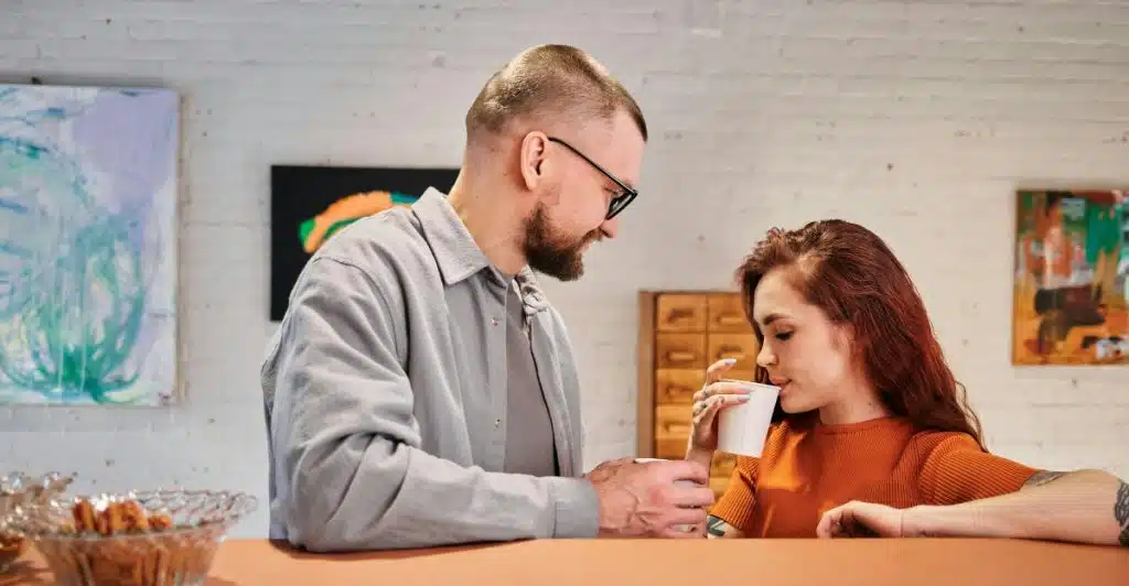 A man with a beard and glasses stands looking down at a woman with red hair who is sipping from a white cup, as they sit at a counter in a modern space with abstract art on the wall behind them.