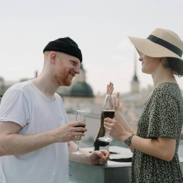 A man and a woman on a rooftop, holding glasses of red wine and smiling at each other, with the city skyline in the background under an overcast sky