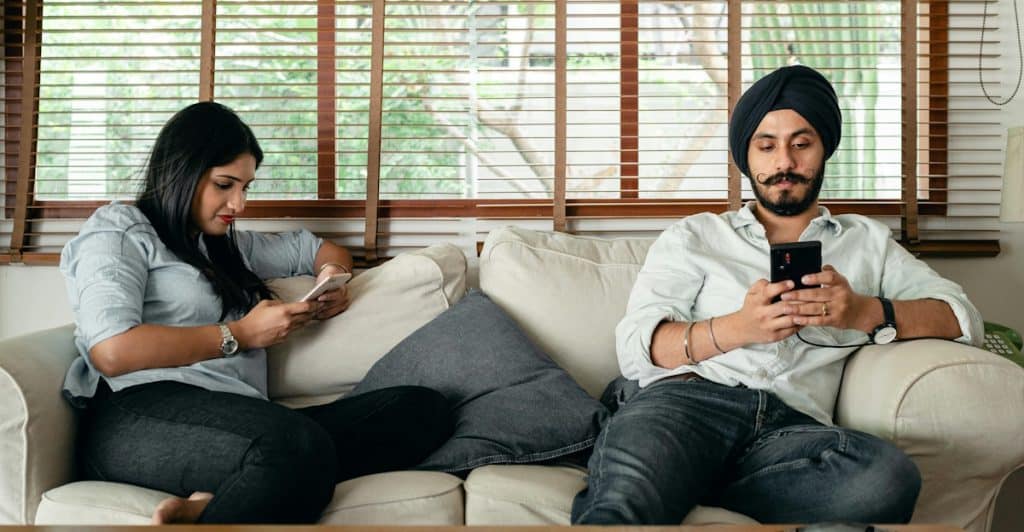 A man and a woman are seated on a light-colored couch in a living room, both looking at their individual smartphones, with the woman on the left wearing glasses and the man on the right wearing a black turban.