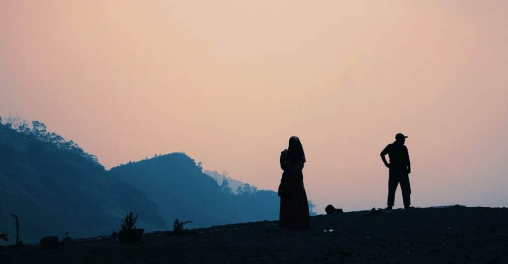 A man and a woman standing on a hill at sunset, with mountains in the background.