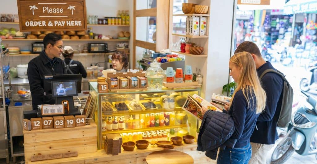 A man and a woman standing at a counter in a bright cafe, looking at a menu, with a person behind the counter.