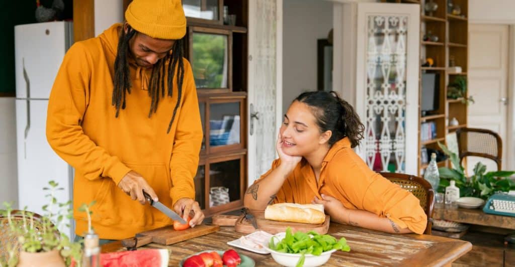 A man chopping food on a cutting board while a woman watches him in a bright kitchen