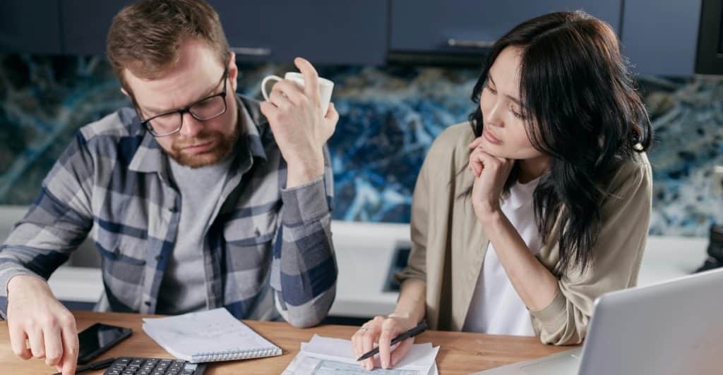 A man and a woman sitting at a table with a laptop and papers, both looking at the documents, with the man holding a mug.