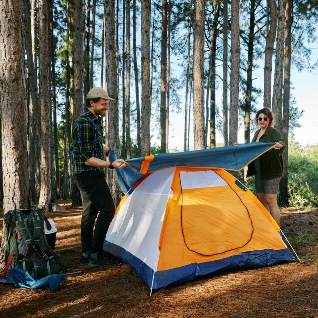  man and a woman setting up a colorful orange and blue tent in a pine forest, with backpacks nearby