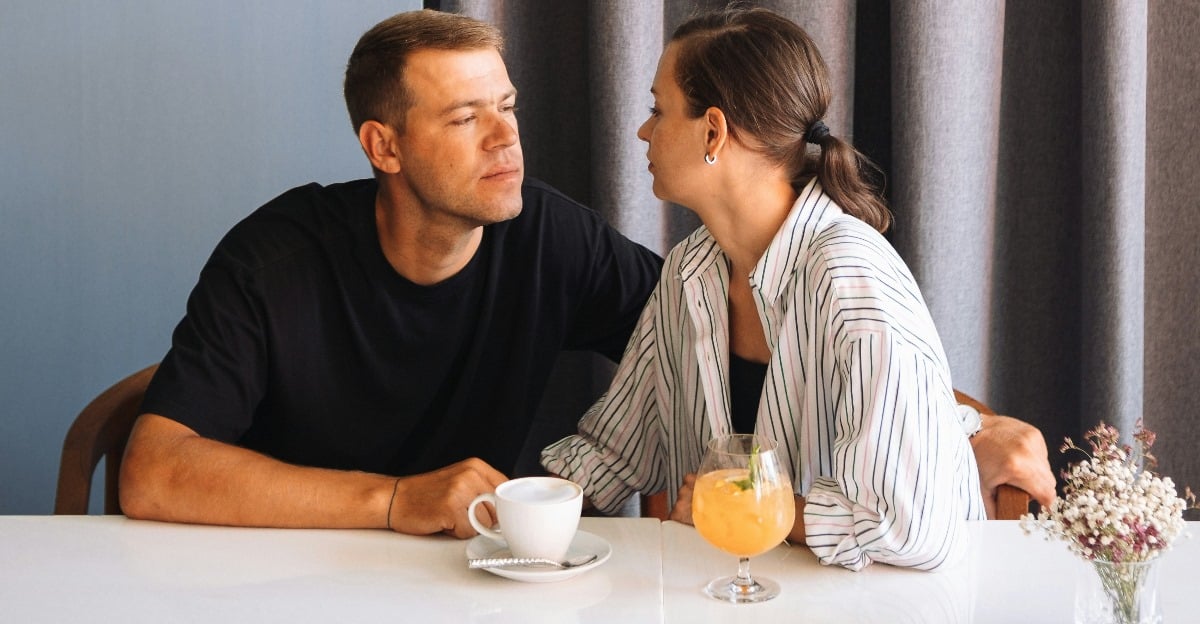 A man and woman sitting at a table looking into each other’s eyes with drinks in front of them and curtains in the background
