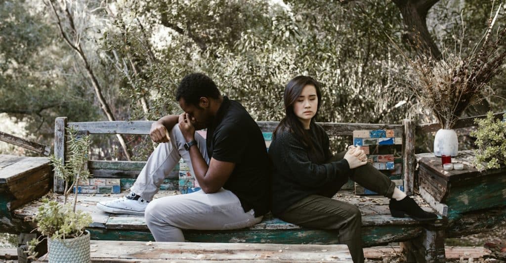 A man and a woman sitting back-to-back on a wooden bench outdoors looking upset and in distress.