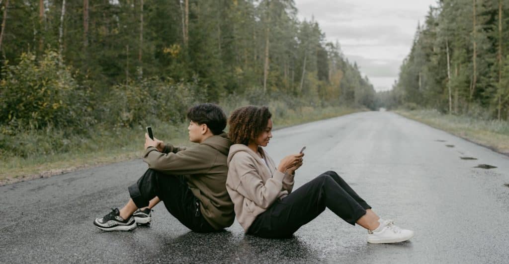 A man and a woman sitting on a wet road, leaning back-to-back while looking at their phones.