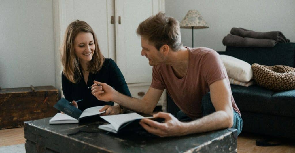 A man and a woman sitting at a coffee table in a living room, both with open notebooks, talking and smiling.