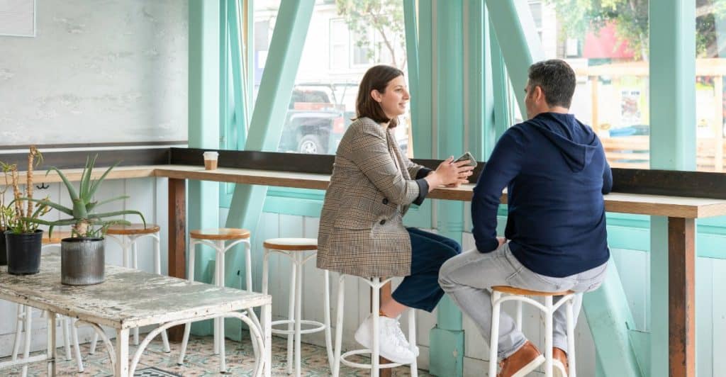 A man and a woman sitting at a counter in a cafe, facing each other and talking, with large windows in the background.