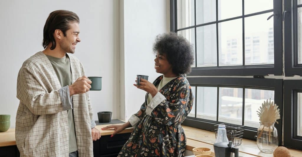 A smiling man in a patterned robe is standing in a kitchen next to a woman with curly hair who is wearing an orange shirt, and both are holding mugs and looking at each other, with a breakfast spread on the counter in front of them.