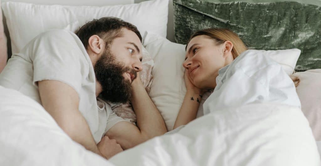 A man and a woman lying in bed, looking at each other and smiling.