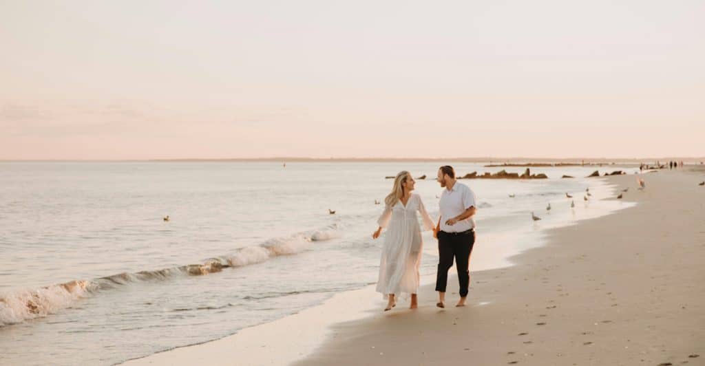 A couple walking barefoot on a sandy beach near the water at sunset.
