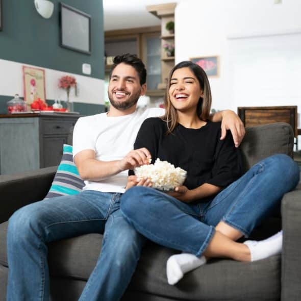 A smiling couple sitting on a couch, watching a movie and eating popcorn together.