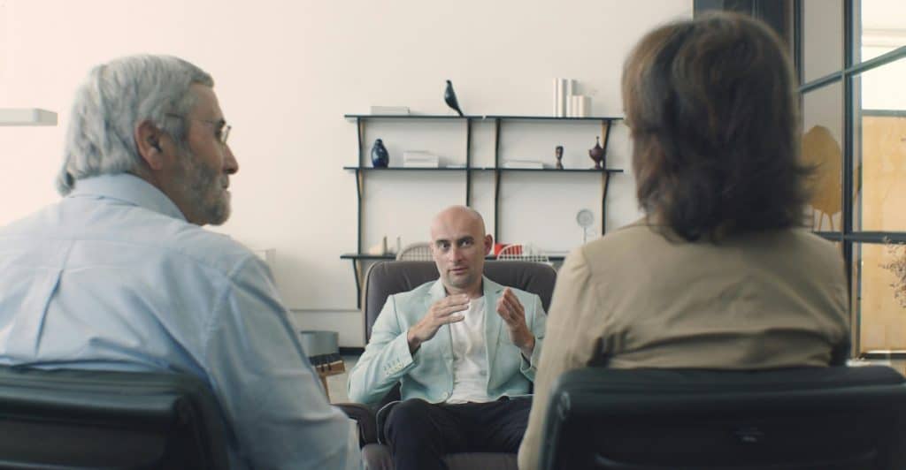 An older couple, seen from behind, sitting in chairs and looking forward at a male counselor who is gesturing with his hands while speaking to them.