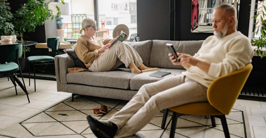 A man sitting in a yellow armchair using his phone and a woman sitting on a gray sofa reading a book, both in a modern living room.