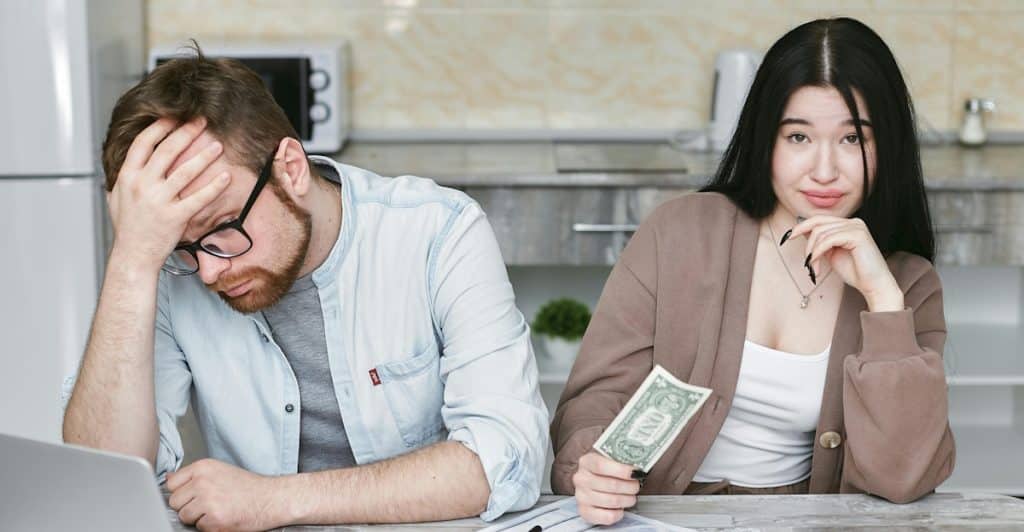 A man looking stressed while looking at a laptop and papers, with a woman holding money next to him.