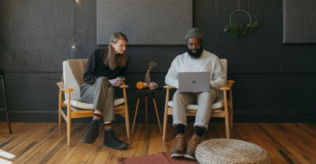 A man sitting with a laptop on his lap while a woman sits next to him in a room with dark walls and wooden floors