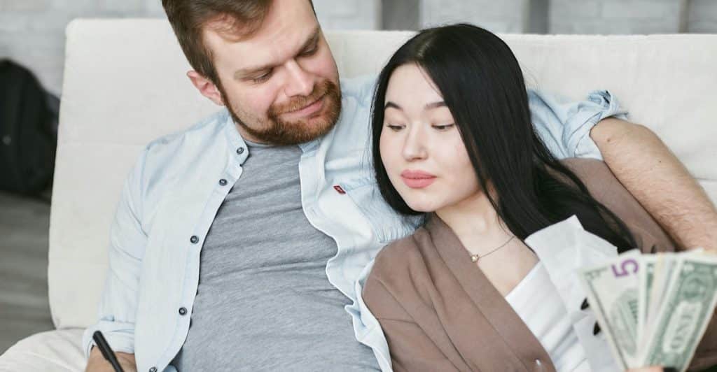 A man and a woman sitting on a sofa, looking at money and a notebook.