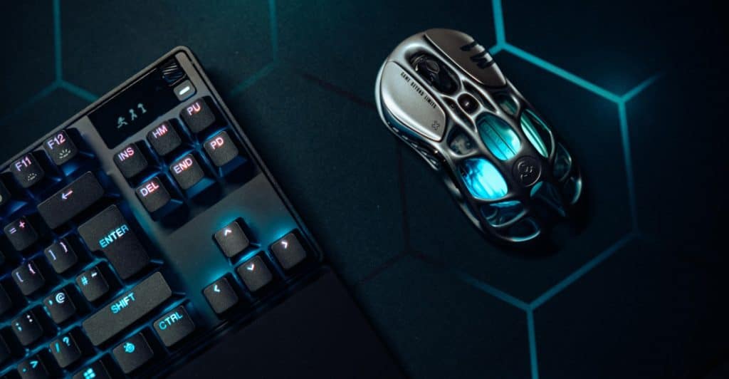 A close-up view of a mechanical keyboard with blue backlighting and a futuristic-looking gaming mouse with green internal lights on a dark mat.