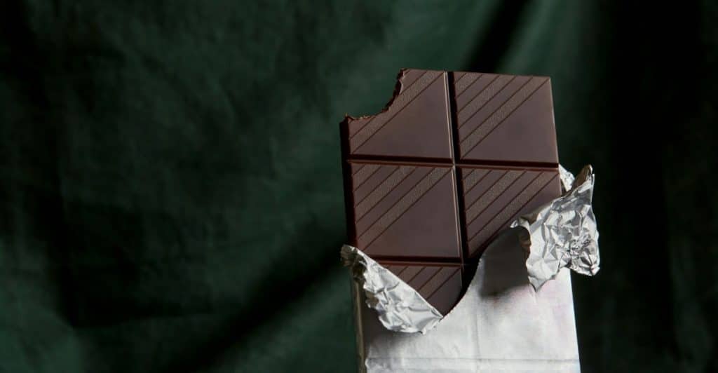 A dark chocolate bar, with one bite taken out, partially unwrapped from its foil on a dark green background.