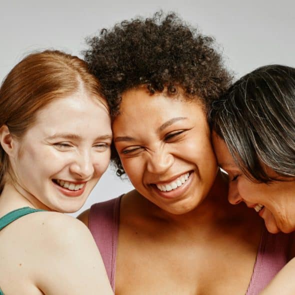 Three women of diverse ethnicities smiling and embracing.